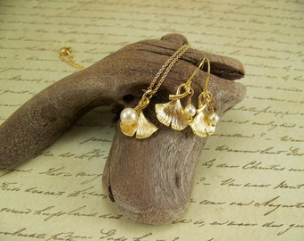 May include: A gold necklace and earrings set featuring ginkgo leaf pendants with a pearl accent. The jewelry is displayed on a piece of driftwood.