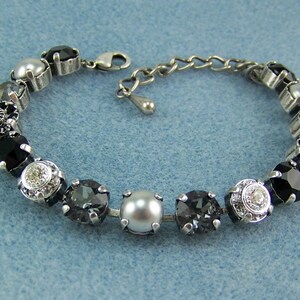 Puede incluir: Una pulsera de tono plateado que presenta una serie de cristales redondos facetados en negro, gris oscuro y transparente, alternados con perlas blancas lisas. La pieza est&aacute; construida con engastes de metal e incluye un cierre de mosquet&oacute;n.