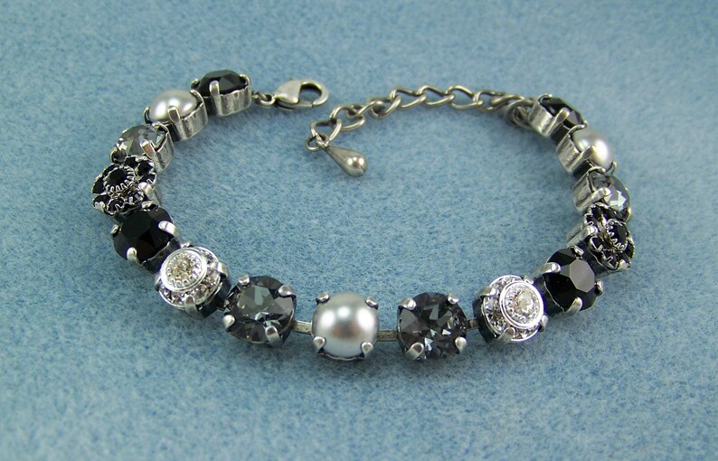 Puede incluir: Una pulsera de tono plateado que presenta una serie de cristales redondos facetados en negro, gris oscuro y transparente, alternados con perlas blancas lisas. La pieza est&aacute; construida con engastes de metal e incluye un cierre de mosquet&oacute;n.