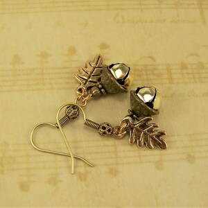May include: A pair of gold tone earrings with acorn and leaf charms. The acorns are silver and gold and have a bell-like shape.