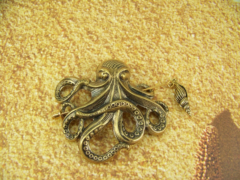 K&ouml;nnte beinhalten: Eine goldfarbene Brosche in Form eines Kraken mit einem detaillierten Design und einem Anh&auml;nger in Form einer Muschel.