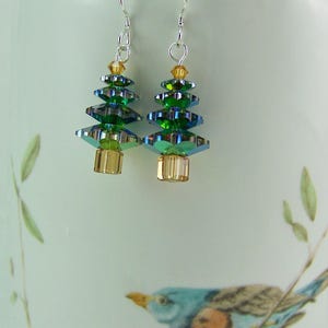 May include: A pair of Christmas tree earrings. Each earring features a layered design of green and iridescent crystals, with a gold-colored base and top. The earrings hang from silver-colored hooks.