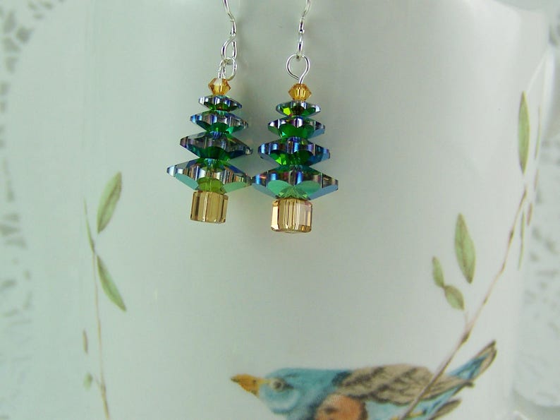 May include: A pair of Christmas tree earrings. Each earring features a layered design of green and iridescent crystals, with a gold-colored base and top. The earrings hang from silver-colored hooks.