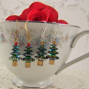 May include: Four Christmas tree earrings with gold, silver, and gold-tone hooks. The trees are made of iridescent green and blue crystals with gold-colored bases and red or amber accents. The earrings are displayed in a floral teacup.