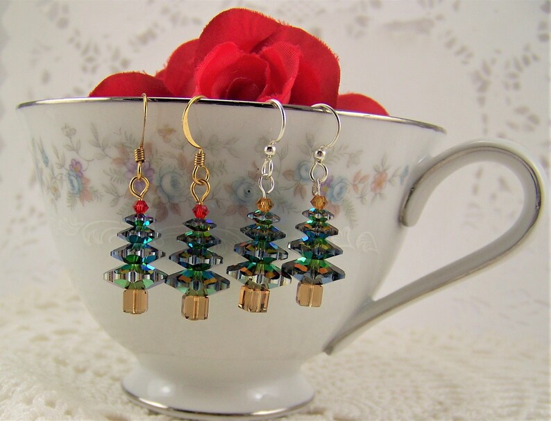May include: Four Christmas tree earrings with gold, silver, and gold-tone hooks. The trees are made of iridescent green and blue crystals with gold-colored bases and red or amber accents. The earrings are displayed in a floral teacup.