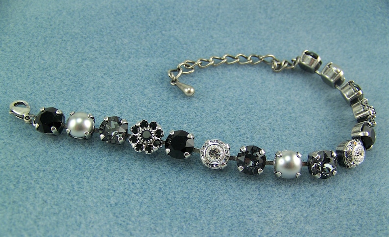 Puede incluir: Una pulsera de tono plateado con una secuencia de cristales negros redondos, perlas grises y piedras facetadas transparentes engastadas en metal. La pulsera incluye una cadena de extensi&oacute;n con un peque&ntilde;o dije en forma de gota y un cierre de mosquet&oacute;n.