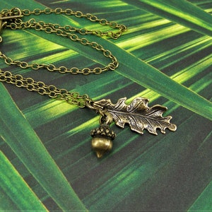 May include: A bronze necklace with an acorn and oak leaf pendant. The necklace is on a green background.