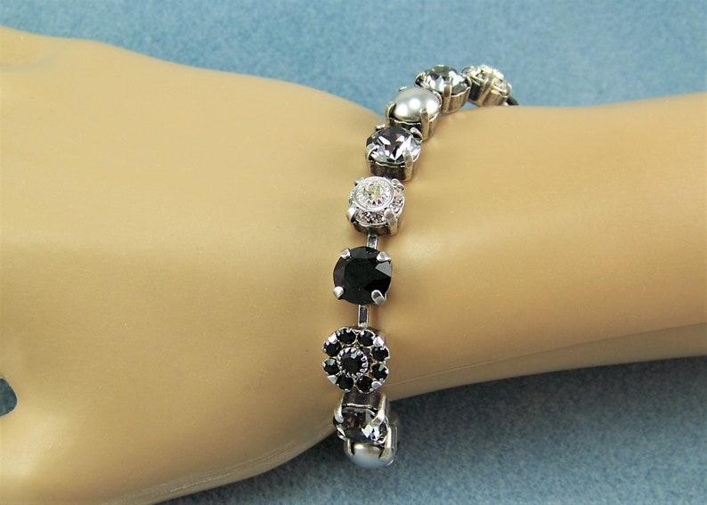 Puede incluir: Una pulsera de tono plateado que presenta una serie de elementos circulares alternos, incluyendo cristales facetados transparentes, piedras negras s&oacute;lidas, una cuenta blanca tipo perla y un peque&ntilde;o racimo floral de piedras negras, engastados.