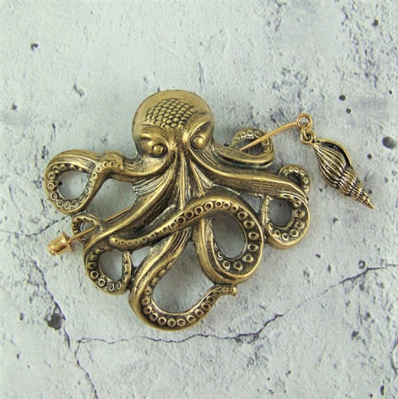 K&ouml;nnte beinhalten: Eine goldfarbene Brosche in Form eines Kraken mit einem Muschel-Anh&auml;nger, der an der Nadel befestigt ist. Der Krake hat ein detailliertes Design mit strukturierten Tentakeln.