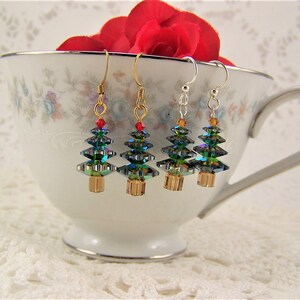 May include: Four Christmas tree earrings with gold, silver, and gold-tone hooks. The earrings feature iridescent green and blue crystal tree shapes with red and orange accents. The earrings are displayed in a white teacup with floral patterns.