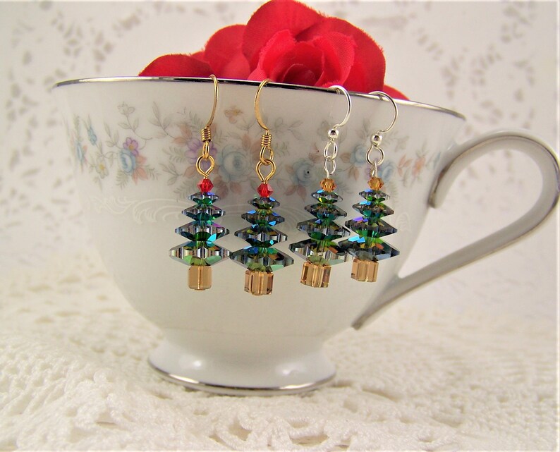 May include: Four Christmas tree earrings with gold, silver, and gold-tone hooks. The earrings feature iridescent green and blue crystal tree shapes with red and orange accents. The earrings are displayed in a white teacup with floral patterns.