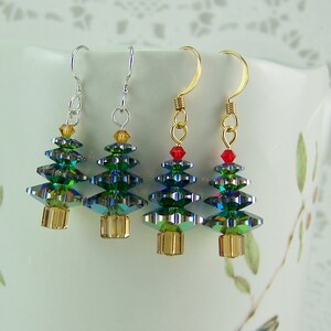 May include: Four Christmas tree earrings with green, blue, and gold crystal beads. The earrings have gold or silver hooks and are displayed against a white background. Each tree has a gold base and a small red or gold bead at the top.