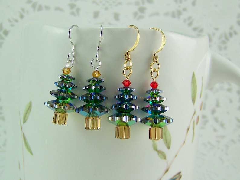 May include: Four Christmas tree earrings with green, blue, and gold crystal beads. The earrings have gold or silver hooks and are displayed against a white background. Each tree has a gold base and a small red or gold bead at the top.