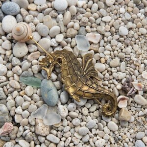 K&ouml;nnte beinhalten: Eine messingfarbene Seepferdchen-Brosche mit strukturiertem, segmentiertem K&ouml;rper und einer langen, d&uuml;nnen Nadel, die nach oben zu einem kleinen, spiralf&ouml;rmigen Muschelaufsatz f&uuml;hrt. Das Seepferdchen hat einen geschwungenen Schwanz und definierte Flossen.