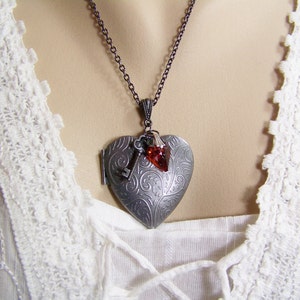May include: A silver heart-shaped locket necklace with a key charm and a red gemstone. The necklace is on a white lace background.