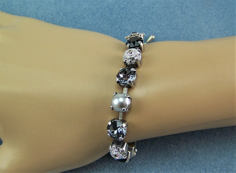 Puede incluir: Una pulsera que presenta una serie de piedras redondas engastadas en tonos variados de negro, gris y plata. El dise&ntilde;o incluye cristales oscuros facetados, una cuenta gris lisa similar a una perla y piedras transparentes en un marco plateado.