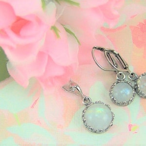 May include: A set of silver earrings and a necklace with iridescent white moonstone cabochons in a silver bezel setting. The jewelry is photographed on a pink and green floral background.