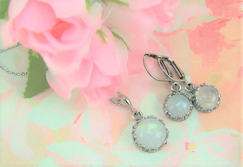 May include: A set of silver earrings and a necklace with iridescent white moonstone cabochons in a silver bezel setting. The jewelry is photographed on a pink and green floral background.
