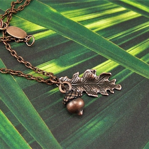 Puede incluir: Un collar de cobre con un colgante de bellota y hoja de roble. El collar está sobre un fondo verde.