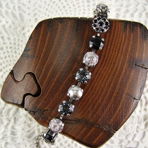 Puede incluir: Una pulsera que presenta una serie de elementos redondos alternos, incluyendo piedras negras facetadas, cristales transparentes y una perla gris lisa, todo engastado en una cadena de metal plateado que sigue una trayectoria curva.