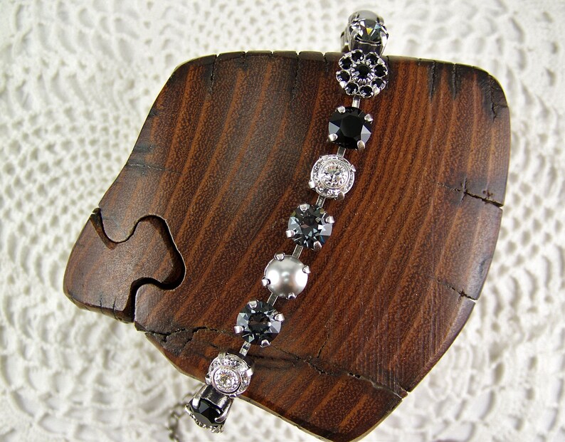 Puede incluir: Una pulsera que presenta una serie de elementos redondos alternos, incluyendo piedras negras facetadas, cristales transparentes y una perla gris lisa, todo engastado en una cadena de metal plateado que sigue una trayectoria curva.