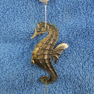 K&ouml;nnte beinhalten: Eine goldfarbene Seepferdchen-Anstecknadel aus Metall mit strukturiertem, segmentiertem K&ouml;rper und einer Flosse. Die Nadel ist mit einer kleinen, runden, wei&szlig;-braunen Spiral-Muschelperle verziert und hat eine lange, d&uuml;nne Metallnadel mit Endkappe.