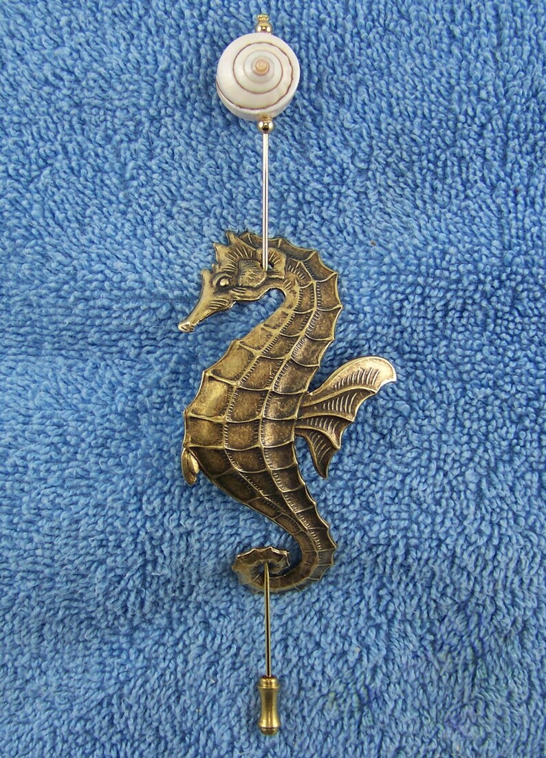 K&ouml;nnte beinhalten: Eine goldfarbene Seepferdchen-Anstecknadel aus Metall mit strukturiertem, segmentiertem K&ouml;rper und einer Flosse. Die Nadel ist mit einer kleinen, runden, wei&szlig;-braunen Spiral-Muschelperle verziert und hat eine lange, d&uuml;nne Metallnadel mit Endkappe.