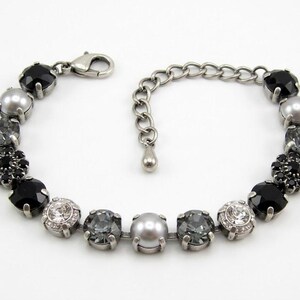 Puede incluir: Una pulsera de tono plateado con una secuencia de cristales redondos facetados en negro, gris y transparente, junto con perlas grises lisas y un grupo de cristales negros en forma de flor. Incluye un cierre de mosquet&oacute;n y una cadena ajustable.
