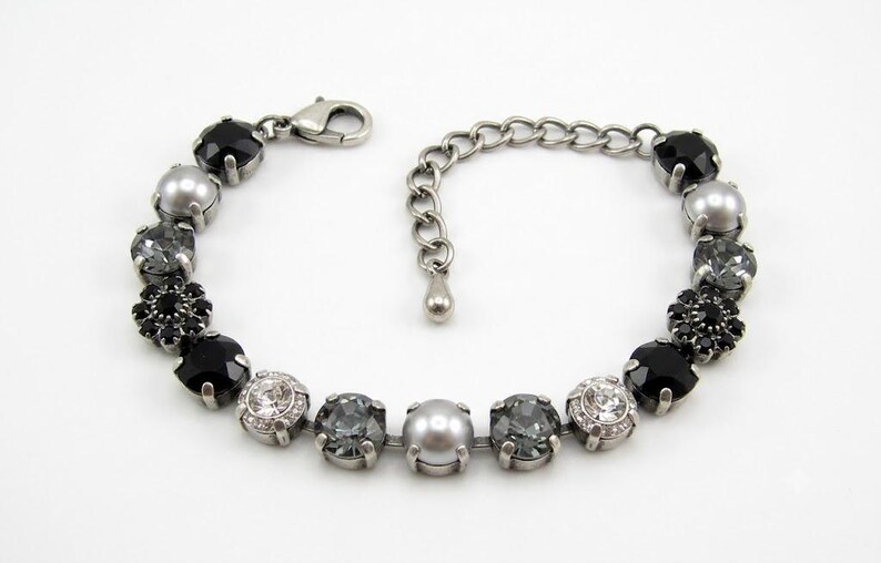 Puede incluir: Una pulsera de tono plateado con una secuencia de cristales redondos facetados en negro, gris y transparente, junto con perlas grises lisas y un grupo de cristales negros en forma de flor. Incluye un cierre de mosquet&oacute;n y una cadena ajustable.