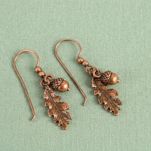 May include: A pair of copper-colored earrings featuring an acorn and oak leaf design. The earrings have a hook closure and are set against a light green background. The acorn and leaf are detailed with intricate textures.