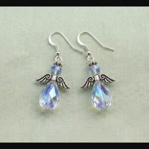 Puede incluir: Un par de pendientes con forma de &aacute;ngel. Cada pendiente presenta un gancho plateado, una peque&ntilde;a cuenta azul y un cristal iridiscente en forma de l&aacute;grima. Los pendientes tambi&eacute;n tienen peque&ntilde;as alas plateadas detalladas. El fondo es verde claro.