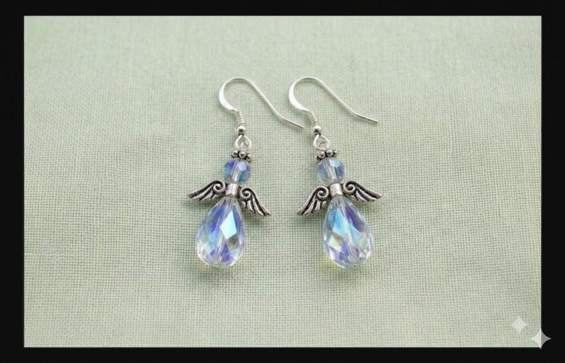 Puede incluir: Un par de pendientes con forma de &aacute;ngel. Cada pendiente presenta un gancho plateado, una peque&ntilde;a cuenta azul y un cristal iridiscente en forma de l&aacute;grima. Los pendientes tambi&eacute;n tienen peque&ntilde;as alas plateadas detalladas. El fondo es verde claro.