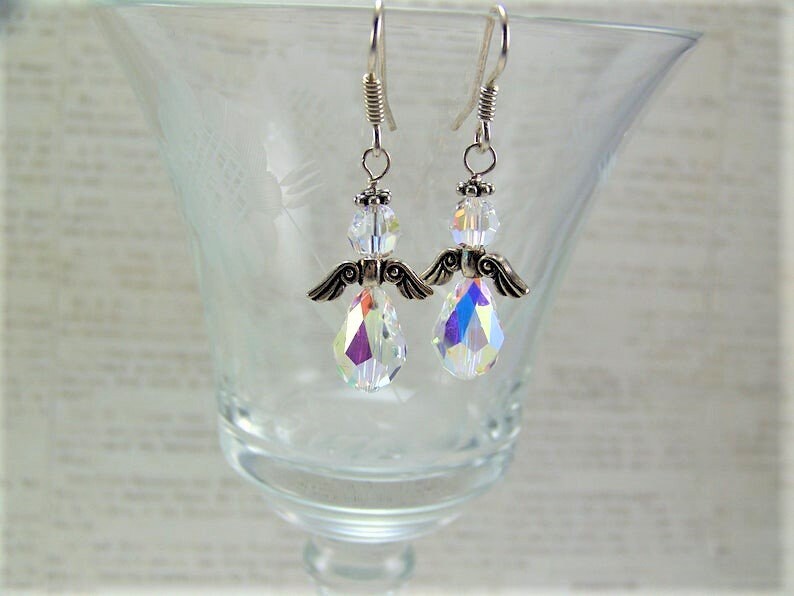 Puede incluir: Un par de pendientes con forma de &aacute;ngel. Cada pendiente presenta un cuerpo de cristal transparente e iridiscente en forma de l&aacute;grima, alas plateadas y una cabeza de cristal transparente. Los pendientes cuelgan de alambres plateados.