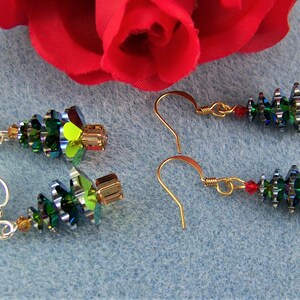 May include: Earrings featuring a Christmas tree design. The earrings are made with green, gold, and red beads, with a gold-colored hook. The earrings are displayed on a light blue surface with a red rose in the background.