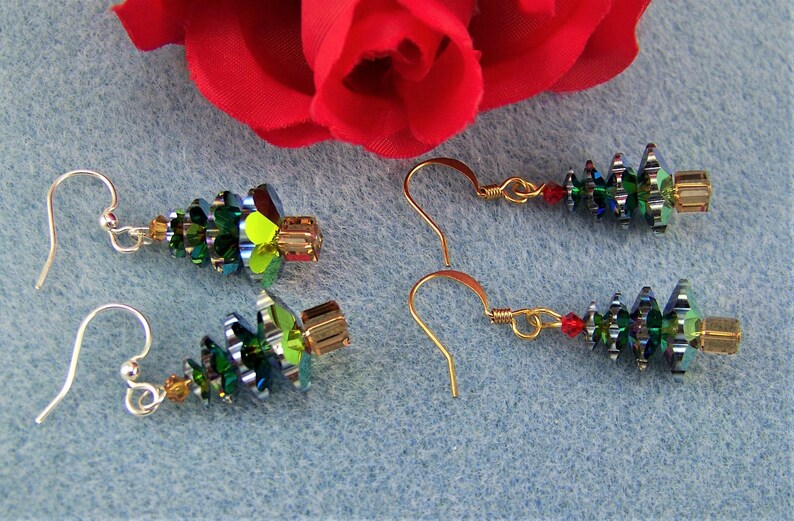 May include: Earrings featuring a Christmas tree design. The earrings are made with green, gold, and red beads, with a gold-colored hook. The earrings are displayed on a light blue surface with a red rose in the background.