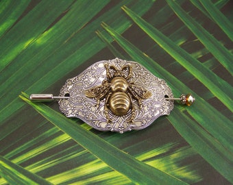 May include: A silver and gold bee hair comb with an ornate design. The comb is on a green background.
