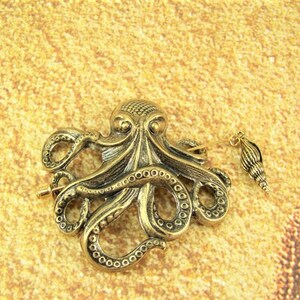 K&ouml;nnte beinhalten: Eine goldfarbene Brosche in Form eines Kraken mit einem Muschel-Anh&auml;nger. Der Krake hat detaillierte Tentakel und einen strukturierten K&ouml;rper.