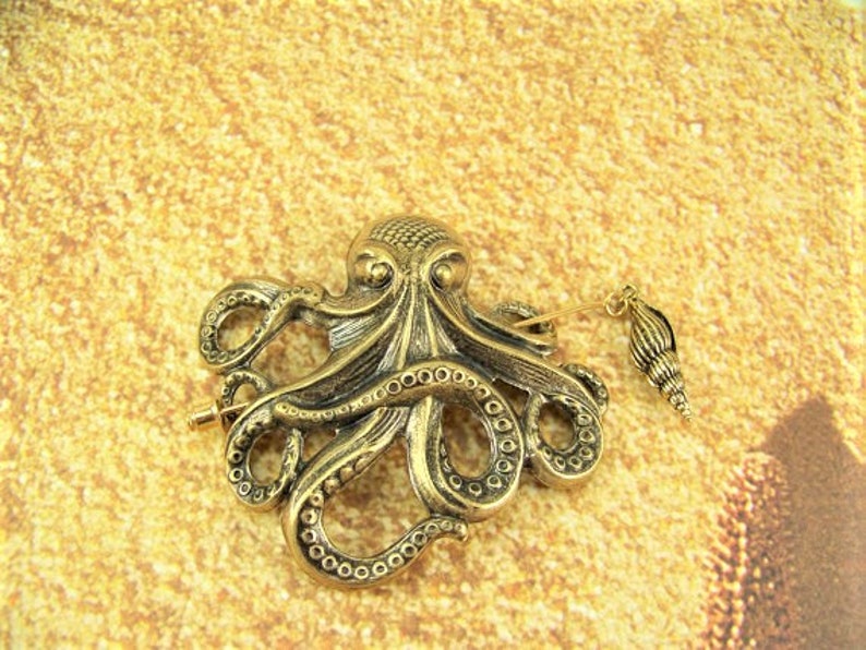 K&ouml;nnte beinhalten: Eine goldfarbene Brosche in Form eines Kraken mit einem Muschel-Anh&auml;nger. Der Krake hat detaillierte Tentakel und einen strukturierten K&ouml;rper.