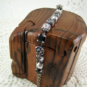Puede incluir: Una pulsera de tono plateado que presenta una secuencia de piedras circulares alternas en negro, transparente y gris, engastadas en garras de metal. La pulsera muestra una variedad de cuentas redondas facetadas y lisas, unidas por una fina cadena de metal plateado.
