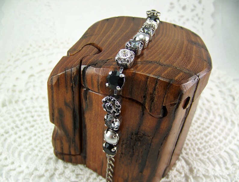 Puede incluir: Una pulsera de tono plateado que presenta una secuencia de piedras circulares alternas en negro, transparente y gris, engastadas en garras de metal. La pulsera muestra una variedad de cuentas redondas facetadas y lisas, unidas por una fina cadena de metal plateado.