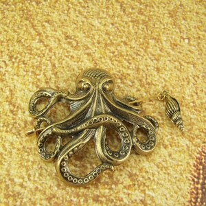 K&ouml;nnte beinhalten: Eine goldfarbene Brosche in Form eines Kraken mit einem Muschel-Anh&auml;nger, der an einer Kette h&auml;ngt. Der Krake hat detaillierte Tentakel und einen strukturierten K&ouml;rper.