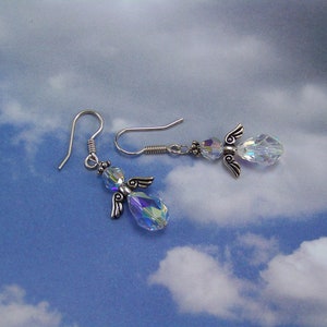 Puede incluir: Un par de pendientes con forma de &aacute;ngel. Cada pendiente presenta un cuerpo de cristal transparente e iridiscente, alas plateadas y un gancho plateado. Los pendientes est&aacute;n colocados sobre un fondo de cielo azul con nubes blancas.