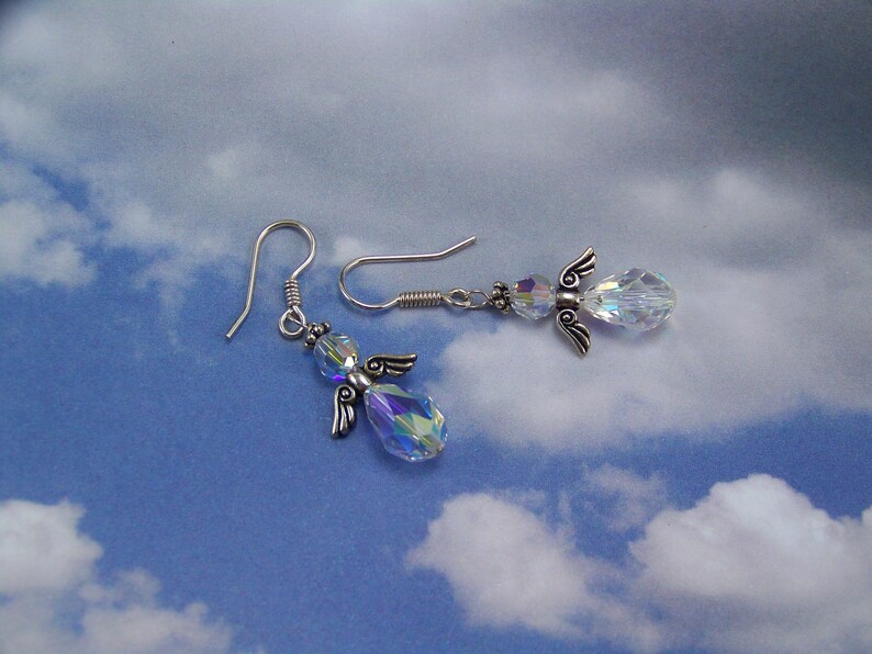 Puede incluir: Un par de pendientes con forma de &aacute;ngel. Cada pendiente presenta un cuerpo de cristal transparente e iridiscente, alas plateadas y un gancho plateado. Los pendientes est&aacute;n colocados sobre un fondo de cielo azul con nubes blancas.
