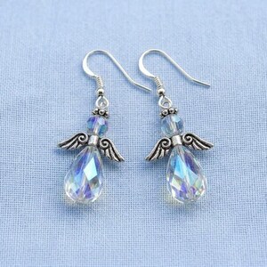 Puede incluir: Un par de pendientes con forma de &aacute;ngel con cristales iridiscentes en forma de l&aacute;grima. Los pendientes tienen alas y ganchos de metal plateado, sobre un fondo de tela azul claro.