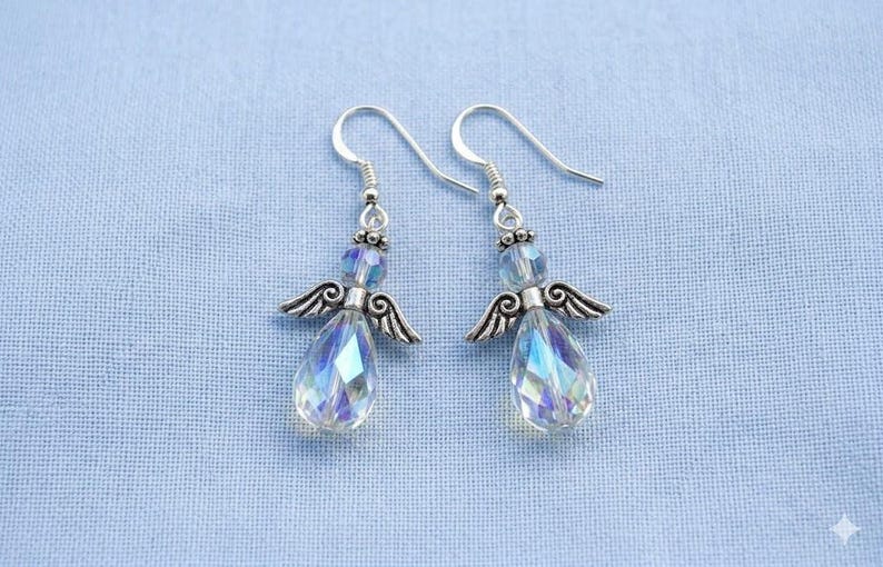 Puede incluir: Un par de pendientes con forma de &aacute;ngel con cristales iridiscentes en forma de l&aacute;grima. Los pendientes tienen alas y ganchos de metal plateado, sobre un fondo de tela azul claro.