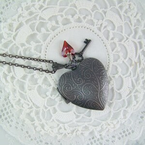 May include: A silver heart-shaped locket necklace with a key charm and a red crystal pendant. The necklace is on a white lace background.