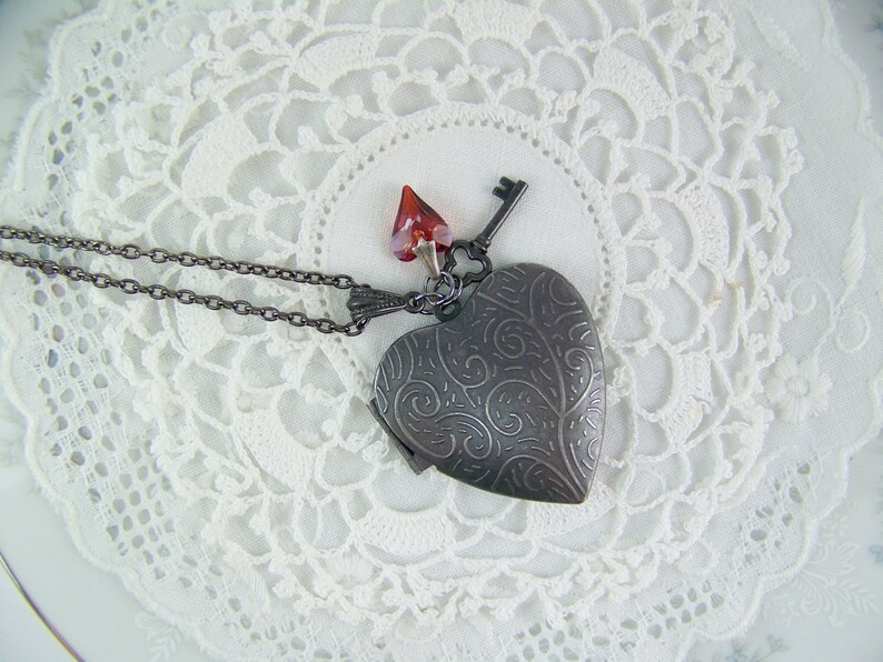 May include: A silver heart-shaped locket necklace with a key charm and a red crystal pendant. The necklace is on a white lace background.