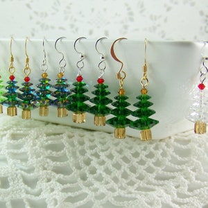 May include: A collection of Christmas tree earrings. The earrings feature green, blue, and clear crystal tree designs with gold-colored bases and red or green bead accents. The earrings are displayed on a white surface.