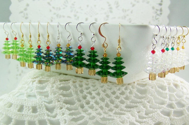 May include: A collection of Christmas tree earrings. The earrings feature green, blue, and clear crystal tree designs with gold-colored bases and red or green bead accents. The earrings are displayed on a white surface.