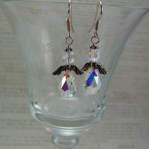 Puede incluir: Un par de pendientes con forma de &aacute;ngel con colgantes de cristal iridiscente en forma de l&aacute;grima. Los pendientes presentan alas y detalles met&aacute;licos plateados, con una cuenta de cristal transparente en la parte superior. Los pendientes cuelgan de ganchos plateados.
